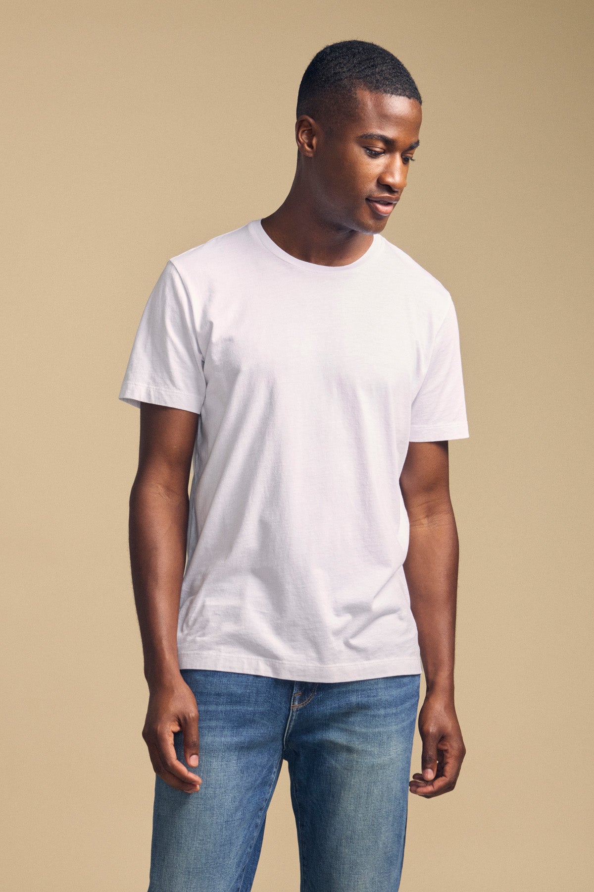 A man stands against a beige background wearing the Velvet by Graham & Spencer HOWARD TEE, a plain white lightweight Pima jersey t-shirt, paired with blue jeans. He looks downward with his hands relaxed by his sides.