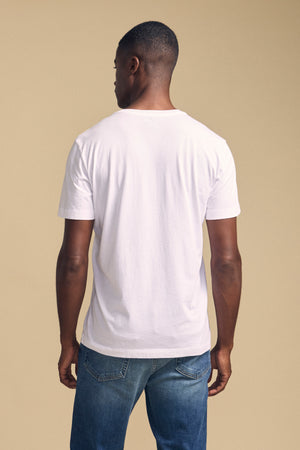 A man stands with his back to the camera against a beige background, wearing blue jeans and the Velvet by Graham & Spencer HOWARD TEE, a plain white lightweight Pima jersey T-shirt.