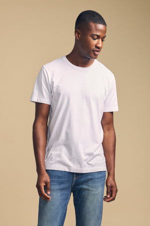 A man stands against a beige background wearing the Velvet by Graham & Spencer HOWARD TEE, a plain white lightweight Pima jersey t-shirt, paired with blue jeans. He looks downward with his hands relaxed by his sides.
