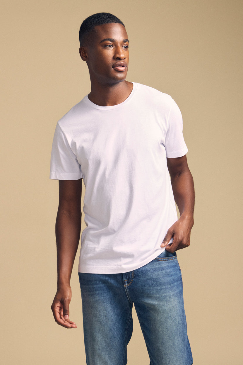 A man in the HOWARD TEE by Velvet by Graham & Spencer, a plain white Pima jersey shirt, and blue jeans stands against a beige background, looking to the side with one hand by his waist, highlighting this vintage-inspired essential’s perfect fit.