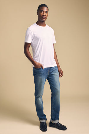 A man stands against a beige background wearing the HOWARD TEE by Velvet by Graham & Spencer—a plain white Pima jersey t-shirt—paired with blue jeans and black shoes for a classic, vintage feel.