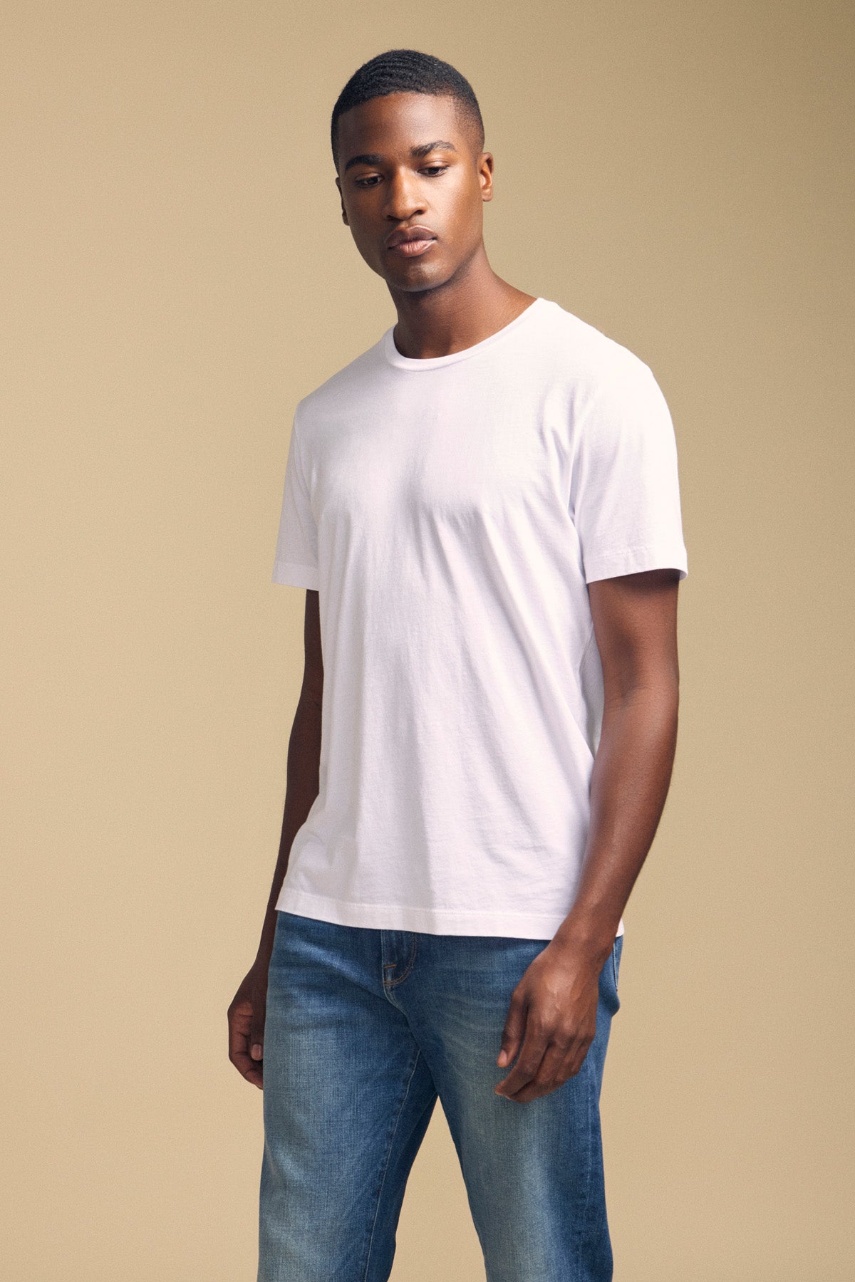 A man wearing the HOWARD TEE by Velvet by Graham & Spencer, a plain white Pima jersey t-shirt with a perfect fit, and blue jeans stands against a beige background.