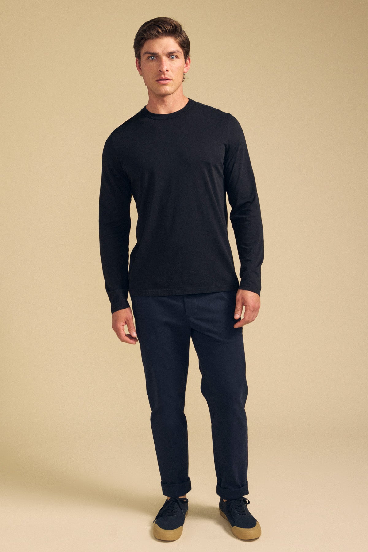 A man stands facing forward wearing the Velvet by Graham & Spencer ROWLAND TEE, a long-sleeve black shirt with a classic crew neckline, paired with dark pants and black sneakers with tan soles against a beige background.-41769768419521