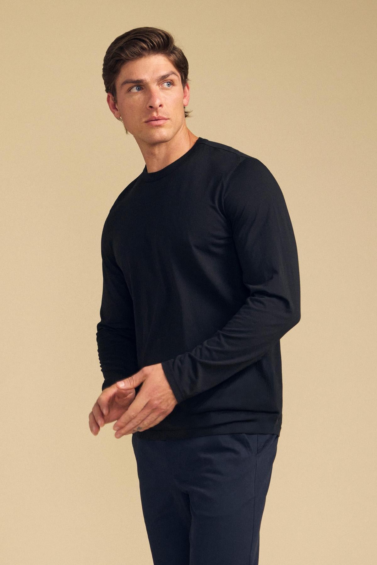 A man stands against a beige background wearing the ROWLAND TEE by Velvet by Graham & Spencer—a black Pima jersey long-sleeve tee featuring a crew neckline—paired with dark pants. He looks slightly to his left with his hands loosely clasped.