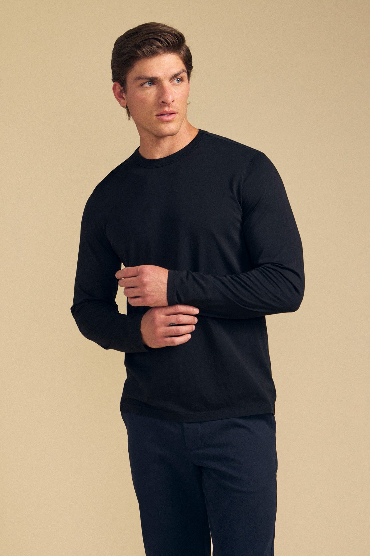 A person wearing the ROWLAND TEE by Velvet by Graham & Spencer—a long-sleeve black top with a classic crew neckline—paired with dark pants stands against a plain beige background, looking to the side with arms slightly bent.