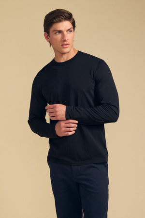 A person wearing the ROWLAND TEE by Velvet by Graham & Spencer—a long-sleeve black top with a classic crew neckline—paired with dark pants stands against a plain beige background, looking to the side with arms slightly bent.