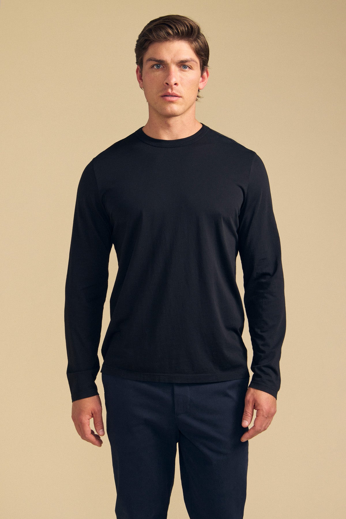 A man stands facing forward, wearing the ROWLAND TEE by Velvet by Graham & Spencer—a plain black long-sleeve tee with a classic crew neckline—paired with dark pants against a neutral beige background.
