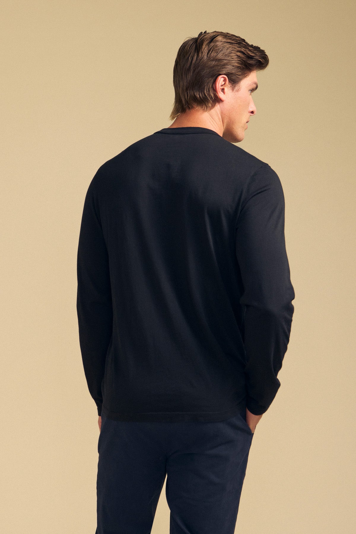 A man with brown hair is shown from the back wearing the ROWLAND TEE by Velvet by Graham & Spencer—a long-sleeve Pima jersey shirt with a classic crew neckline—paired with dark pants, standing against a plain beige background.-41769768321217
