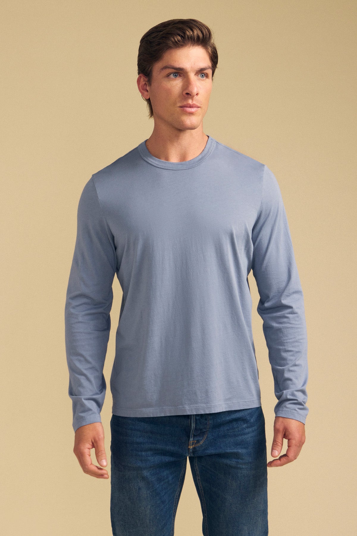 A man wearing the ROWLAND TEE by Velvet by Graham & Spencer—a light blue Pima jersey long-sleeve crewneck—paired with blue jeans stands facing forward against a plain beige background.-41702119112897