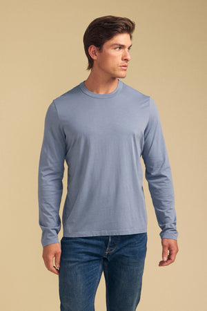 A man wearing the light blue ROWLAND TEE by Velvet by Graham & Spencer and blue jeans stands against a plain beige background, looking to his left.