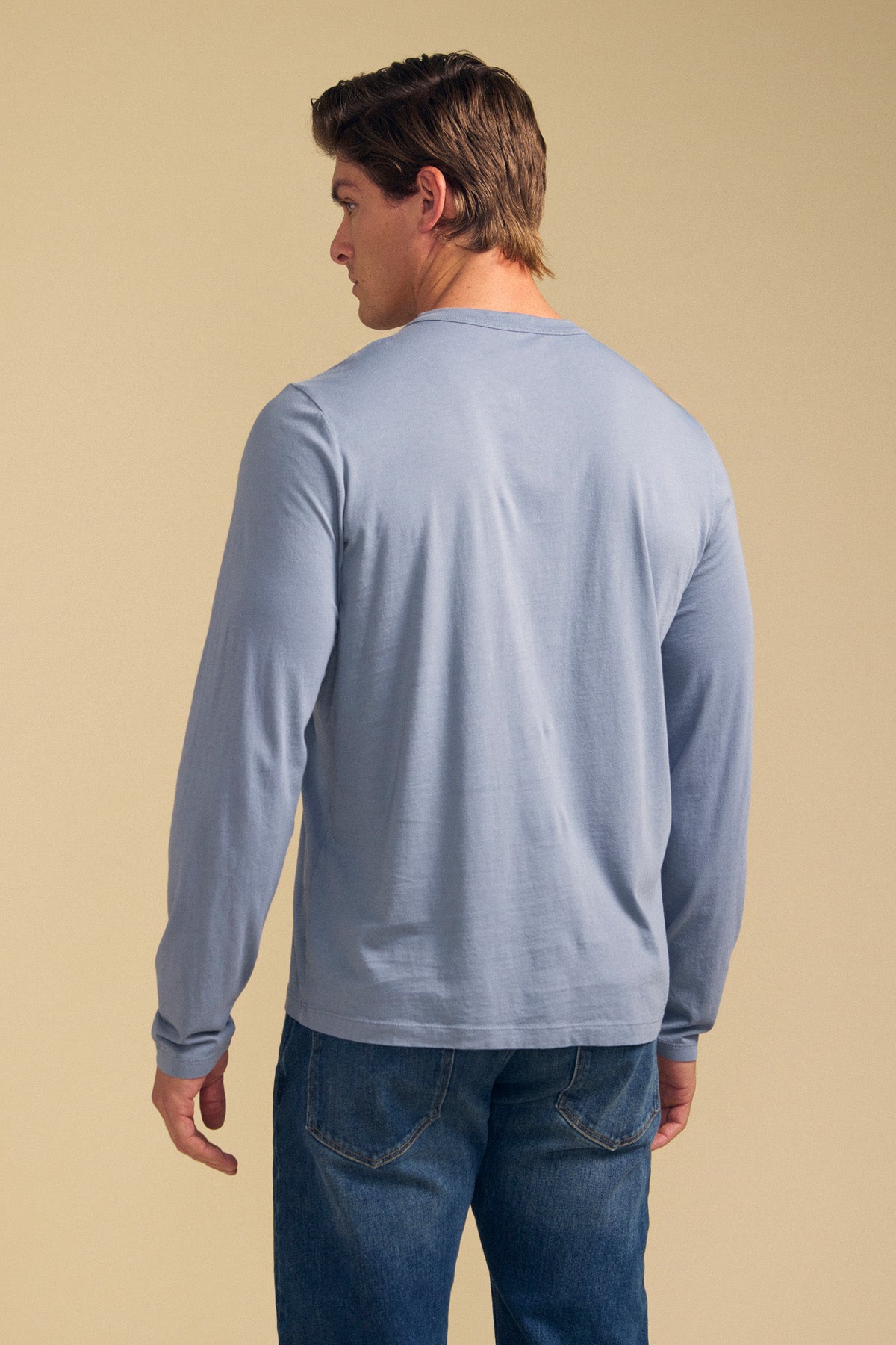 Someone stands with their back to the camera against a beige background, wearing the Velvet by Graham & Spencer ROWLAND TEE—a light blue Pima jersey long-sleeve crewneck—paired with blue jeans.-41702118981825