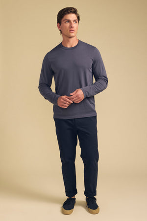 A man stands against a beige background wearing the Velvet by Graham & Spencer ROWLAND TEE, a long-sleeve Pima jersey with a crew neckline, paired with dark pants and black sneakers featuring tan soles.