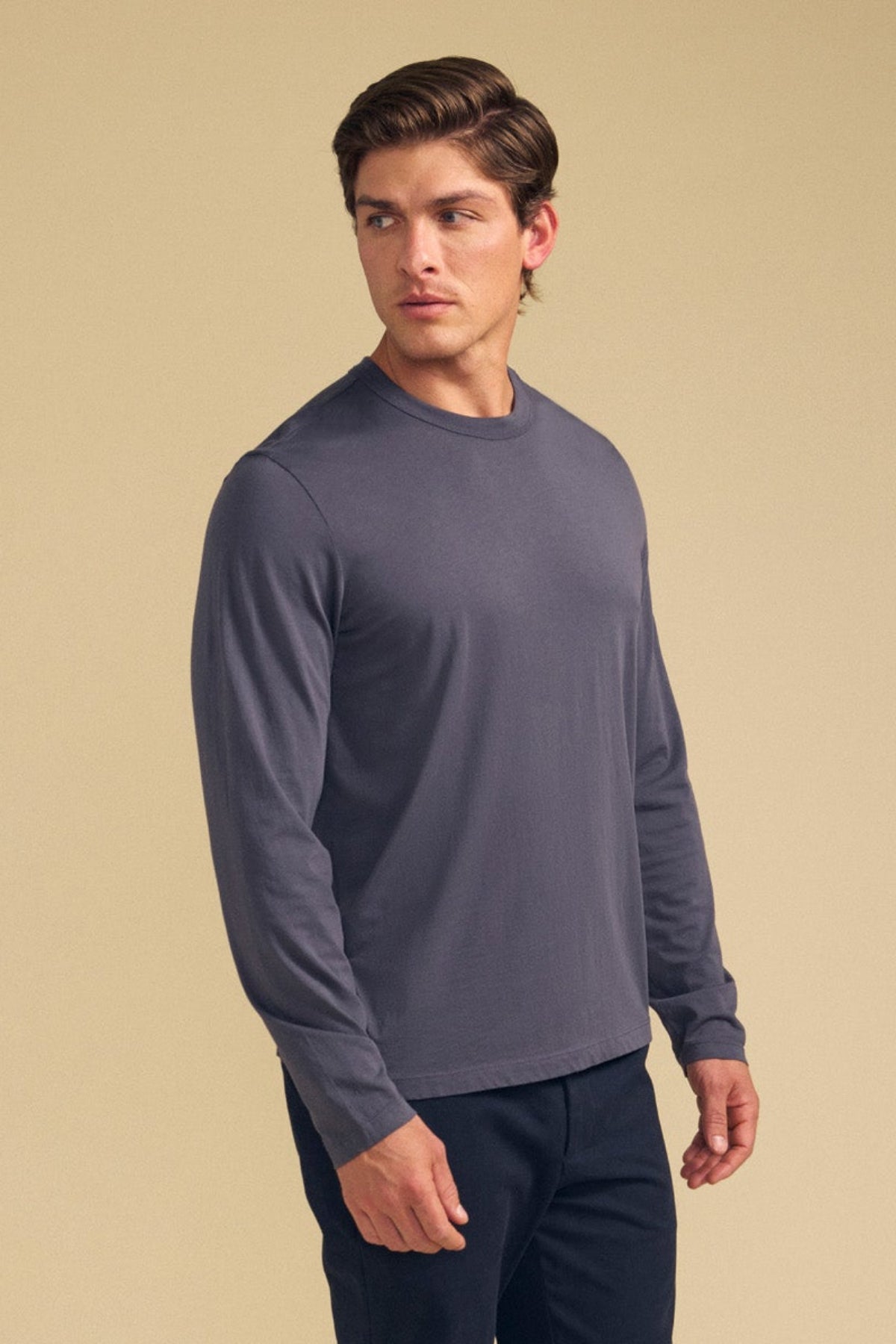 A man wearing the ROWLAND TEE by Velvet by Graham & Spencer, a long-sleeve jersey with a classic crew neckline, and black pants stands against a plain beige background, looking to his left.