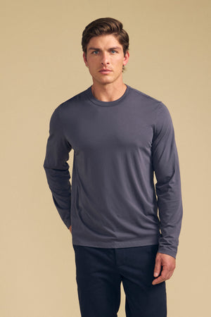 A man faces forward in a Velvet by Graham & Spencer ROWLAND TEE—a grey long-sleeve crew neck—paired with dark pants, set against a plain beige background.