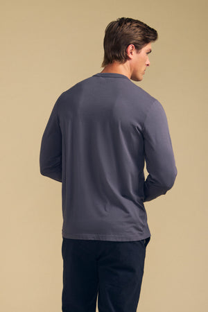 A man faces away from the camera, wearing Velvet by Graham & Spencer’s ROWLAND TEE—a long-sleeve crewneck jersey—and dark pants, set against a plain beige background.