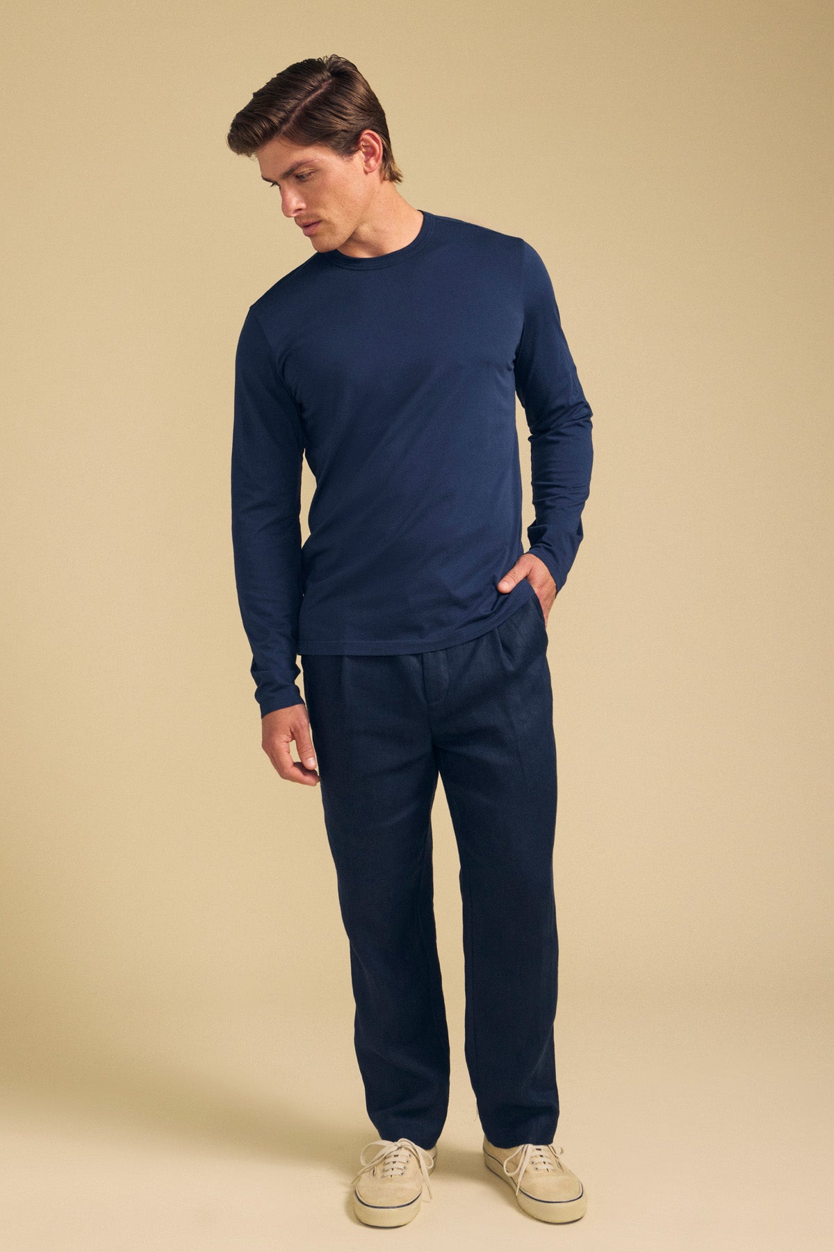 A man stands against a beige background, wearing the ROWLAND TEE by Velvet by Graham & Spencer—a navy long-sleeve crafted from soft Pima jersey with a classic crew neckline—paired with dark pants and light-colored sneakers, one hand in his pocket as he looks down.