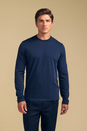 A man with light brown hair wears the Velvet by Graham & Spencer ROWLAND TEE—a plain, long-sleeve navy Pima jersey tee with a classic crew neckline—paired with matching navy pants, standing against a beige background.