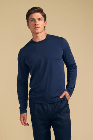 A man wearing the ROWLAND TEE by Velvet by Graham & Spencer—a navy blue, long-sleeve Pima jersey crew neck t-shirt—paired with dark pants stands against a plain beige background, looking forward with one hand in his pocket.