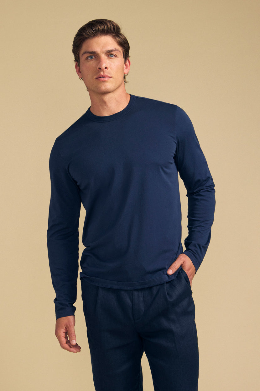 A man wearing the ROWLAND TEE by Velvet by Graham & Spencer—a navy blue, long-sleeve Pima jersey crew neck t-shirt—paired with dark pants stands against a plain beige background, looking forward with one hand in his pocket.