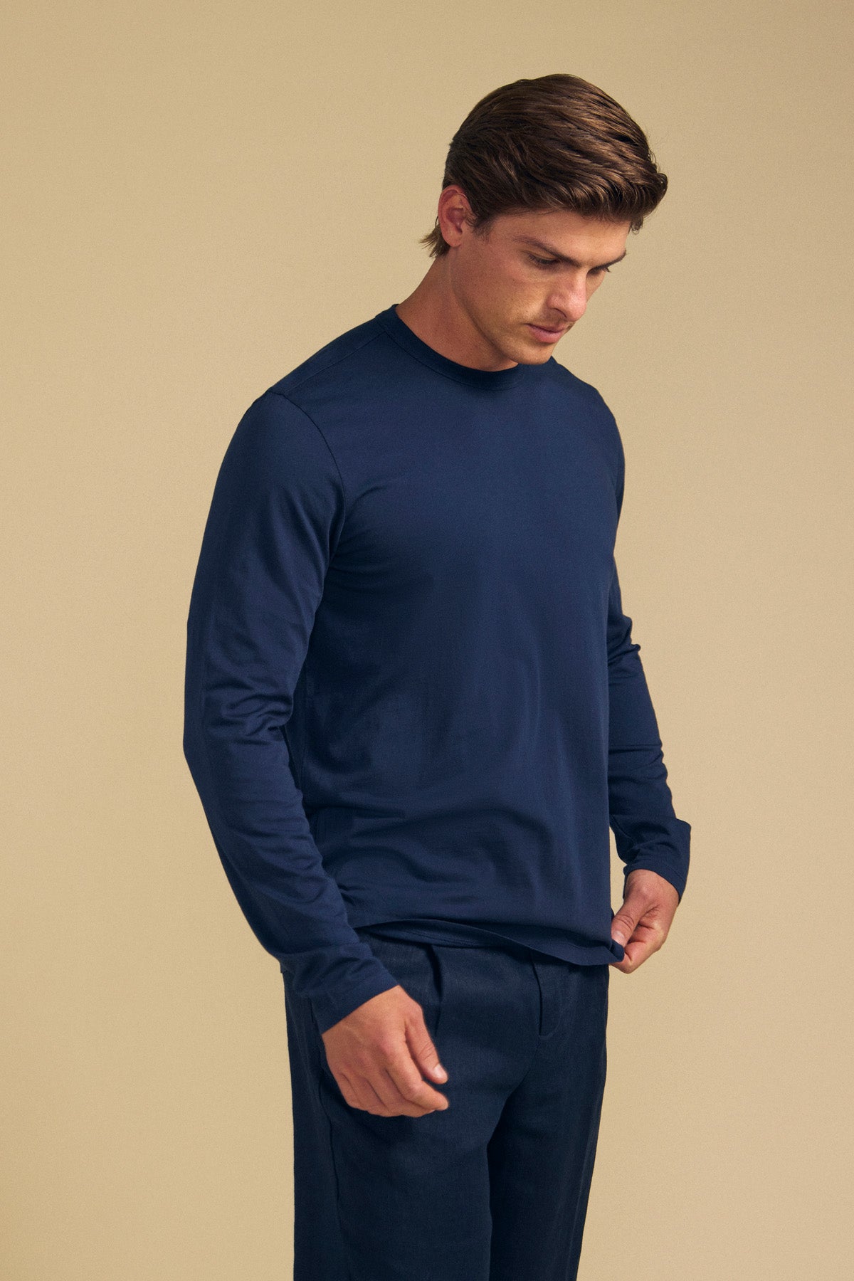 A man stands against a beige background, wearing the Velvet by Graham & Spencer ROWLAND TEE—a navy long-sleeve top crafted from soft Pima jersey with a classic crew neckline—paired with matching navy pants. He gazes slightly downward with his hands by his sides.-41769768190145