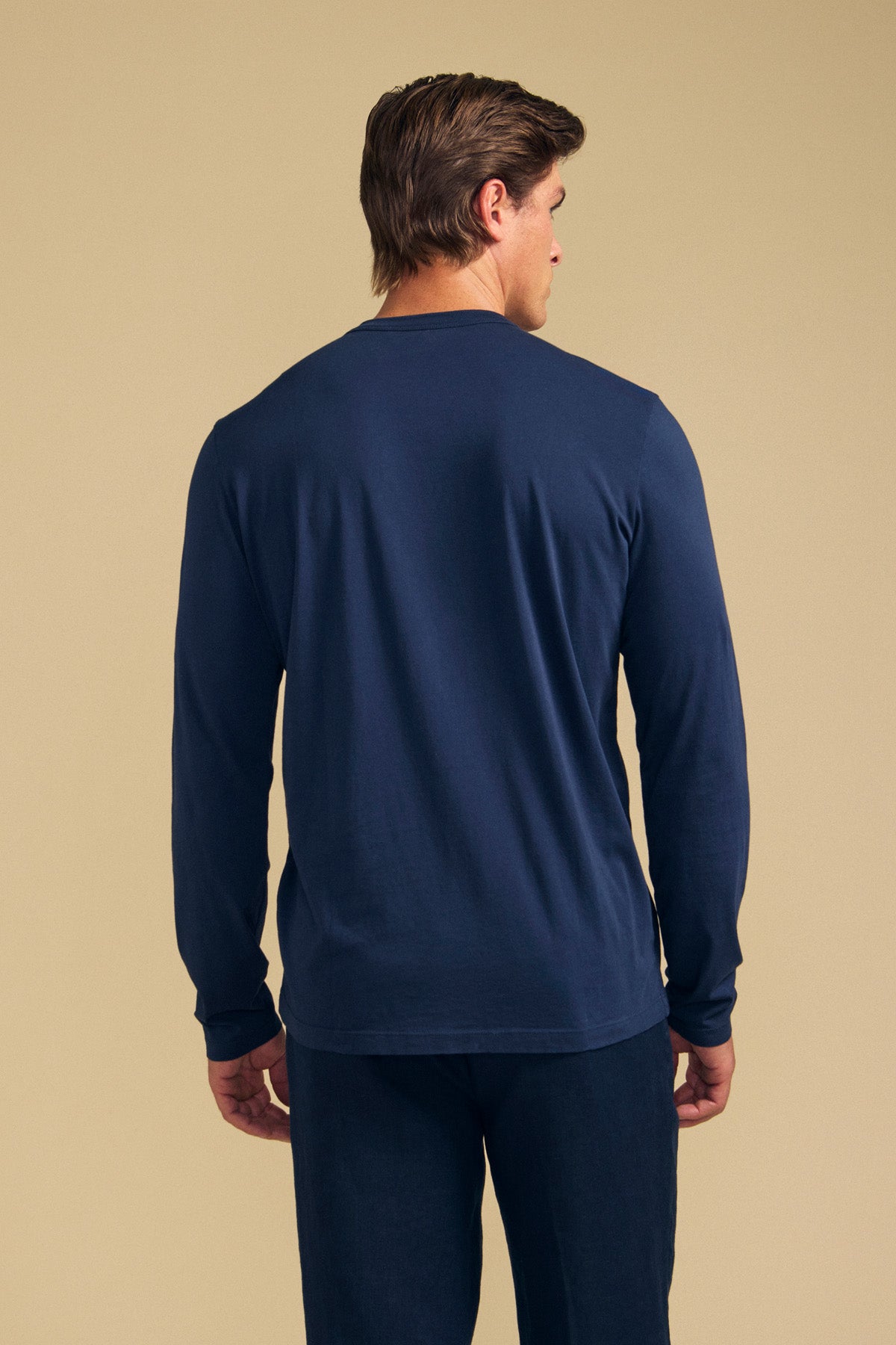 A man is standing with his back to the camera, wearing Velvet by Graham & Spencer's ROWLAND TEE in navy blue. The long-sleeve tee features a classic crew neckline and is paired with matching pants, both crafted from soft Pima jersey, set against a beige background.