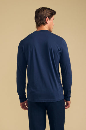 A man is standing with his back to the camera, wearing Velvet by Graham & Spencer's ROWLAND TEE in navy blue. The long-sleeve tee features a classic crew neckline and is paired with matching pants, both crafted from soft Pima jersey, set against a beige background.