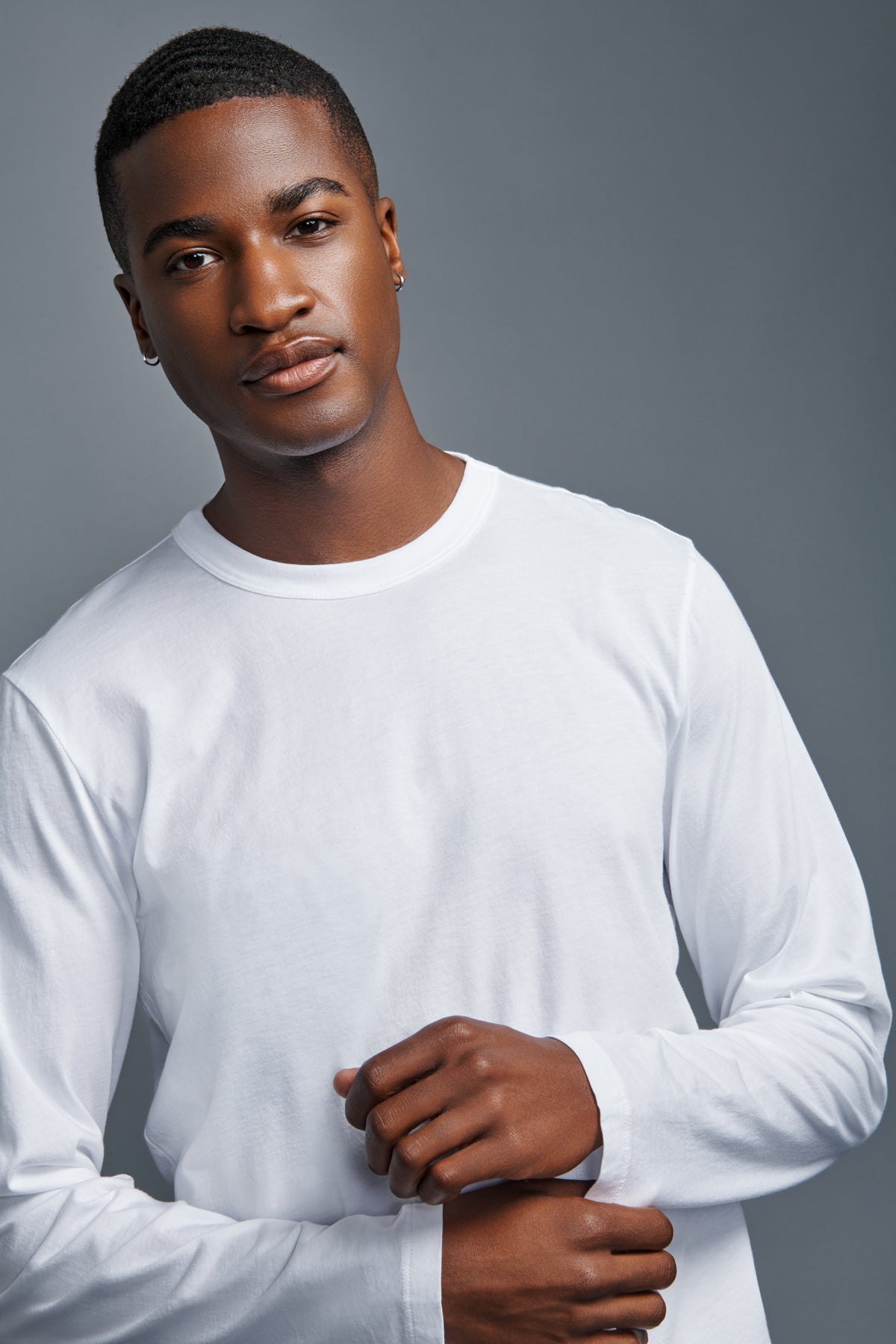 A young man with short hair stands against a gray background, looking at the camera with a neutral expression, wearing the ROWLAND TEE by Velvet by Graham & Spencer—a plain white long-sleeve tee featuring a classic crew neckline.