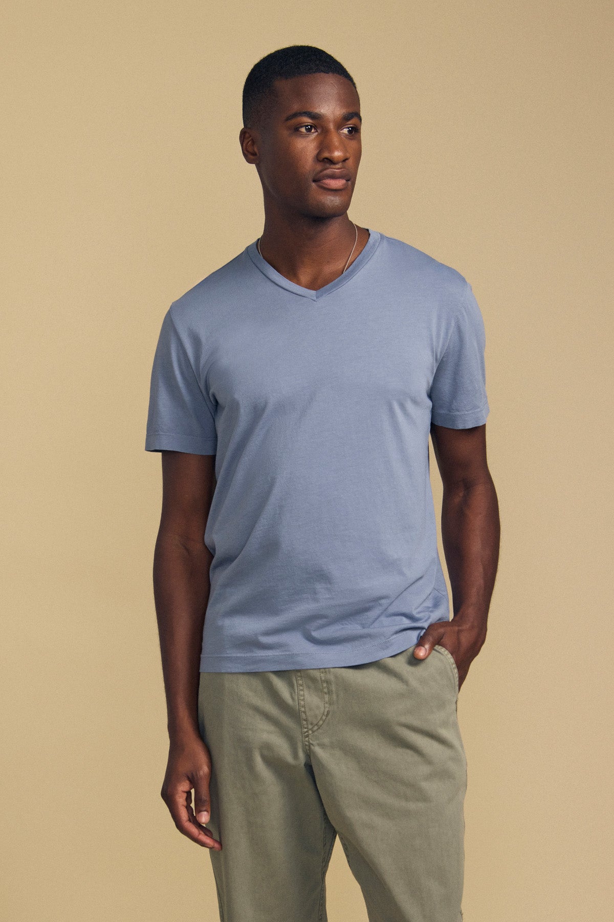 A man stands against a beige background, wearing the SAMSEN TEE by Velvet by Graham & Spencer—a light blue V-neck shirt crafted from soft Pima jersey—paired with light olive pants and one hand in his pocket, making for a stylish and comfortable look for everyday wear.