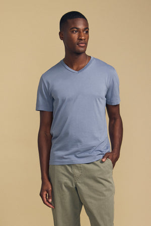 A man stands against a beige background, wearing the SAMSEN TEE by Velvet by Graham & Spencer—a light blue V-neck shirt crafted from soft Pima jersey—paired with light olive pants and one hand in his pocket, making for a stylish and comfortable look for everyday wear.