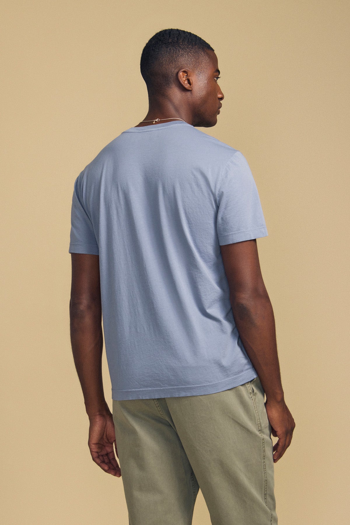 A man stands facing away, wearing the light blue SAMSEN TEE by Velvet by Graham & Spencer paired with light olive pants against a plain beige background—an ideal look for effortless everyday wear.