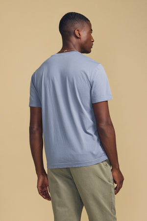 A man stands facing away, wearing the light blue SAMSEN TEE by Velvet by Graham & Spencer paired with light olive pants against a plain beige background—an ideal look for effortless everyday wear.