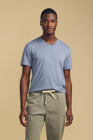 A man stands facing forward against a plain beige background, wearing Velvet by Graham & Spencer’s SAMSEN TEE—a light blue V-neck crafted from soft Pima jersey—paired with light olive-green drawstring pants, creating a look that’s perfect for everyday wear.