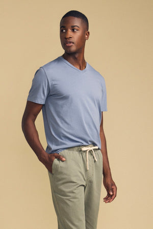 A man stands with one hand in his pocket, wearing the Velvet by Graham & Spencer SAMSEN TEE in light blue paired with light olive drawstring pants, set against a plain beige background—an effortlessly stylish look perfect for everyday wear.