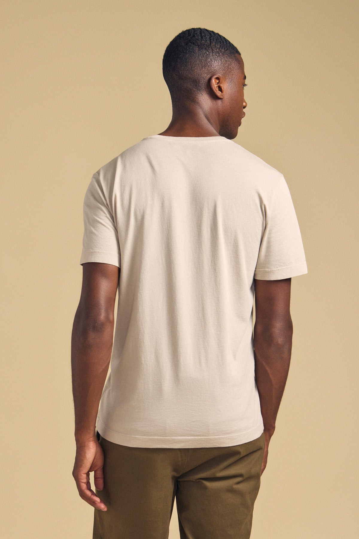 A man stands with his back to the camera, wearing the SAMSEN TEE by Velvet by Graham & Spencer in light beige and olive green pants—ideal for everyday wear—against a neutral background.