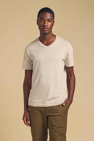 A man stands against a plain beige background wearing the SAMSEN TEE by Velvet by Graham & Spencer, a light beige V-neck Pima jersey tee, paired with olive green pants—ideal for everyday wear.