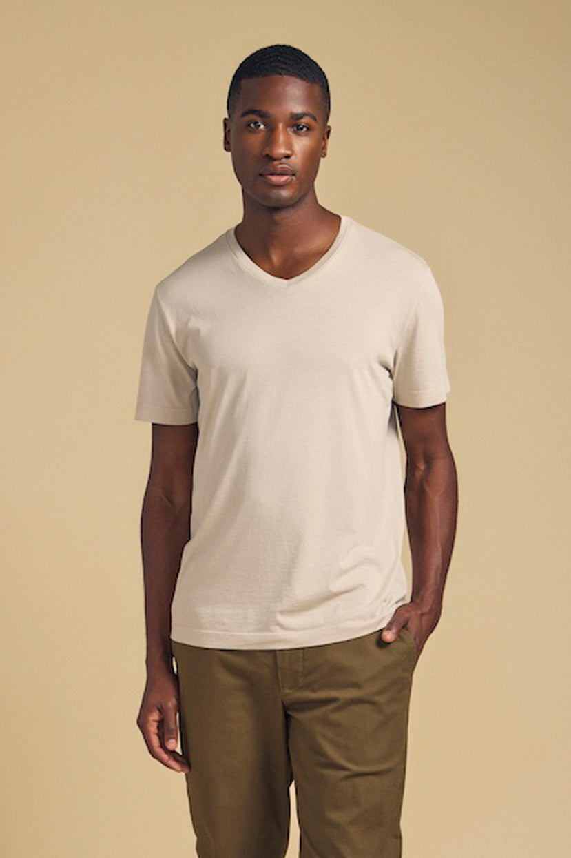 A man stands against a plain beige background wearing the SAMSEN TEE by Velvet by Graham & Spencer, a light beige V-neck Pima jersey tee, paired with olive green pants—ideal for everyday wear.