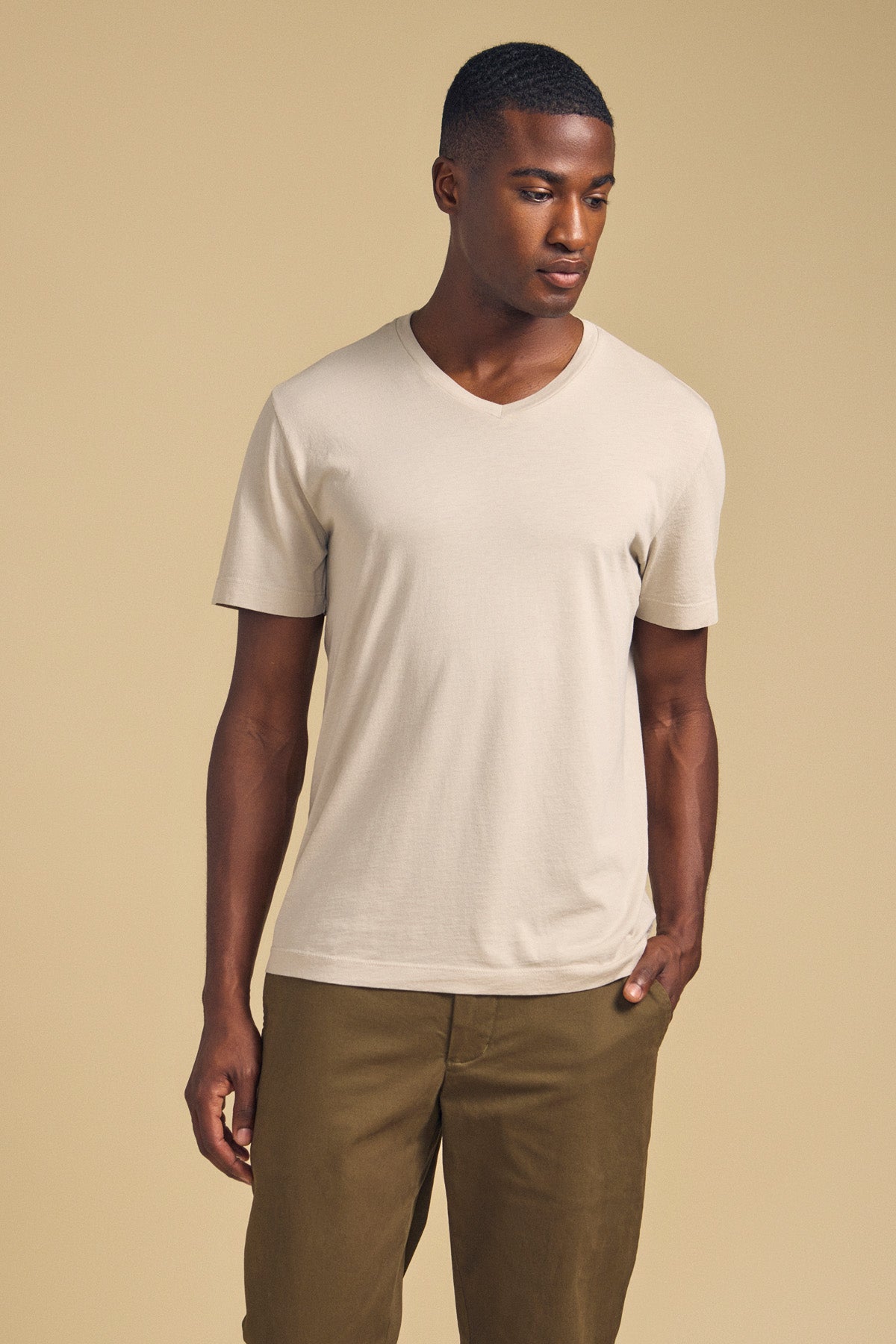 A man wears the SAMSEN TEE by Velvet by Graham & Spencer—a light beige Pima jersey V-neck—paired with olive green pants, standing against a plain beige background for an effortlessly stylish look.