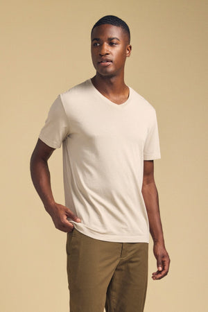 A man stands against a beige background in the SAMSEN TEE by Velvet by Graham & Spencer— a light beige Pima jersey v-neck paired with olive pants, embodying effortless everyday style.
