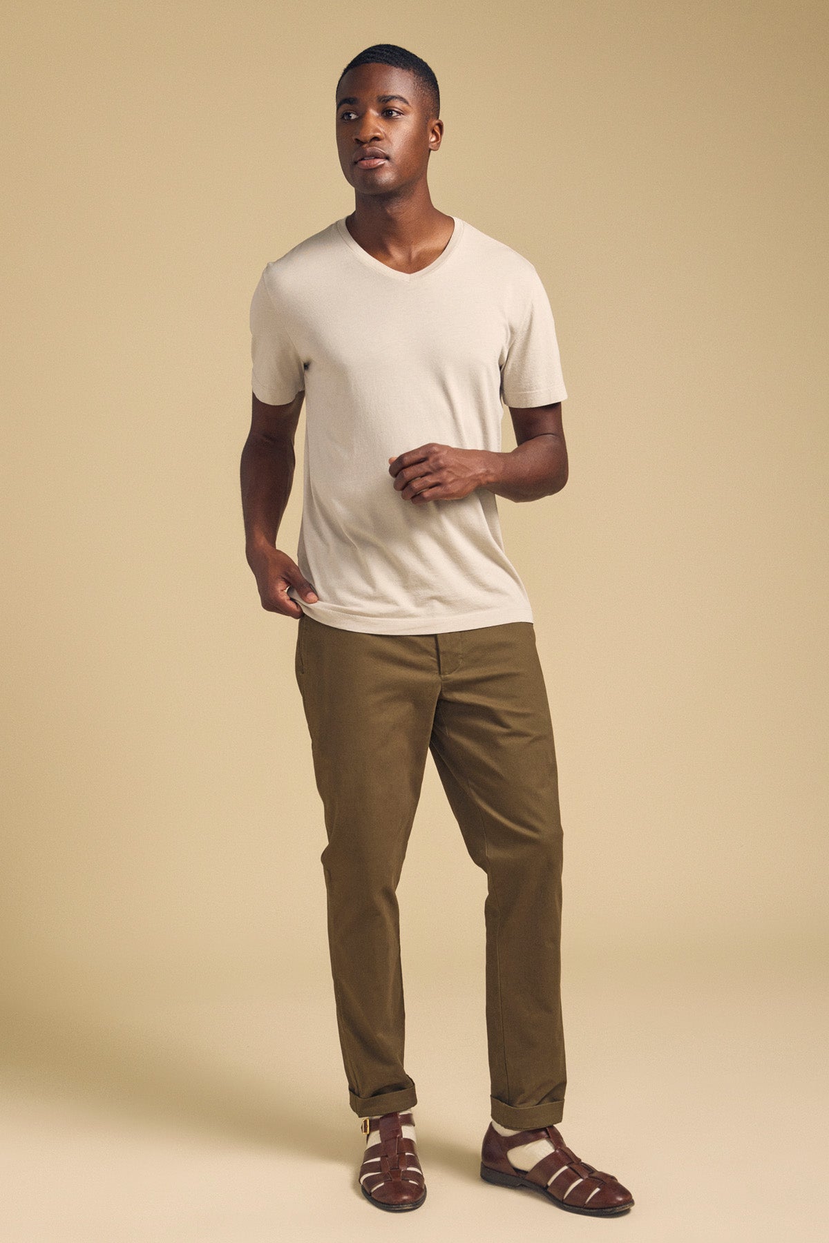 A man stands against a beige background wearing the SAMSEN TEE by Velvet by Graham & Spencer—a light beige Pima jersey V-neck—paired with olive-brown pants and brown sandals, perfect for everyday wear.