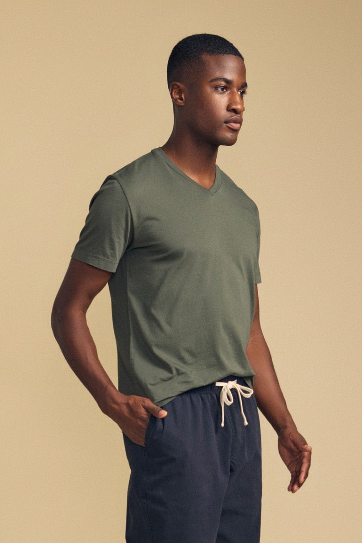 A man stands against a beige background, wearing the SAMSEN TEE by Velvet by Graham & Spencer in olive green with a Pima jersey V-neckline, paired with navy blue drawstring pants and facing slightly to the side—perfect for stylish everyday wear.