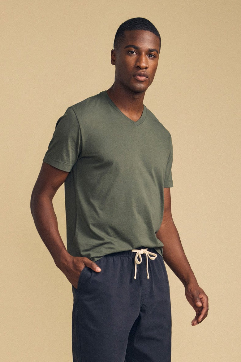 A man wearing the Velvet by Graham & Spencer SAMSEN TEE, a green V-neck top made from soft Pima jersey, pairs it with navy drawstring pants and stands against a beige background with one hand in his pocket, embodying effortless everyday style.