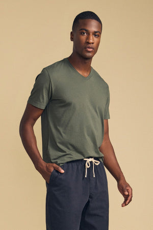 A man wearing the Velvet by Graham & Spencer SAMSEN TEE, a green V-neck top made from soft Pima jersey, pairs it with navy drawstring pants and stands against a beige background with one hand in his pocket, embodying effortless everyday style.