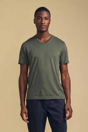 A man stands against a beige background, facing forward with a neutral expression, wearing the Velvet by Graham & Spencer SAMSEN TEE—an olive green V-neck crafted from soft Pima jersey—paired with dark pants; an ideal choice for comfortable everyday wear.