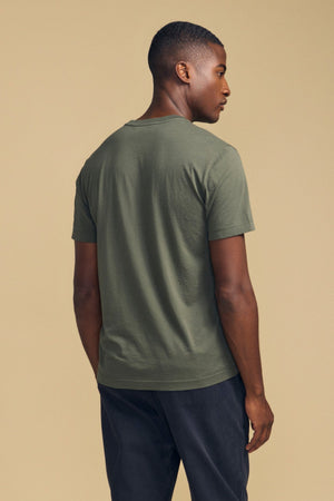 A man stands facing away, wearing the SAMSEN TEE by Velvet by Graham & Spencer in olive green with short sleeves and dark pants, set against a plain beige background—a perfect choice for everyday wear.
