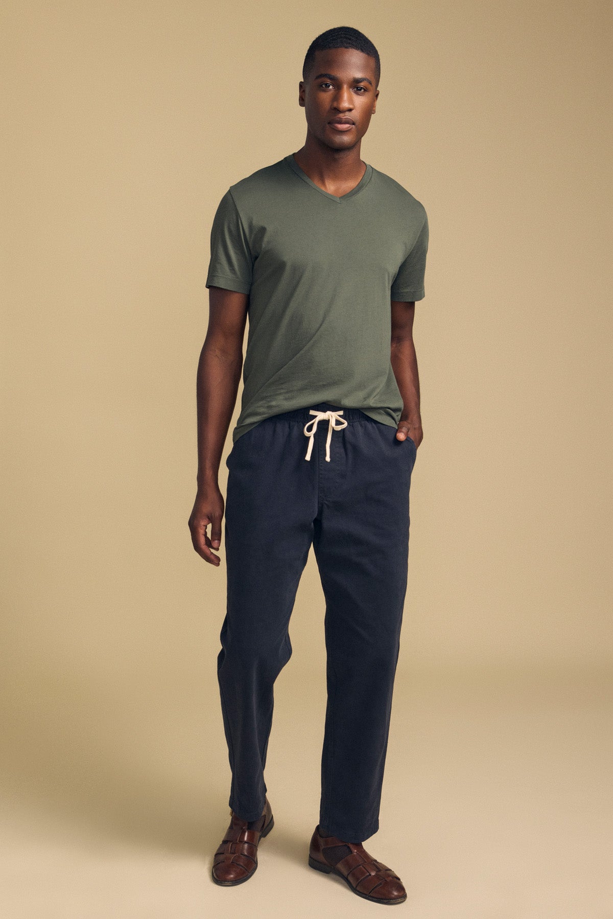 A man stands against a beige background wearing the SAMSEN TEE by Velvet by Graham & Spencer—a green Pima jersey short-sleeve T-shirt—paired with dark drawstring pants and brown shoes for an effortlessly stylish everyday look.-41859803480257