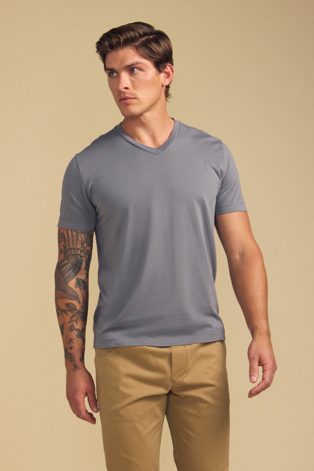 A man with a tattooed left arm wears the SAMSEN TEE by Velvet by Graham & Spencer in gray and khaki pants, standing against a beige background—ideal for everyday wear.