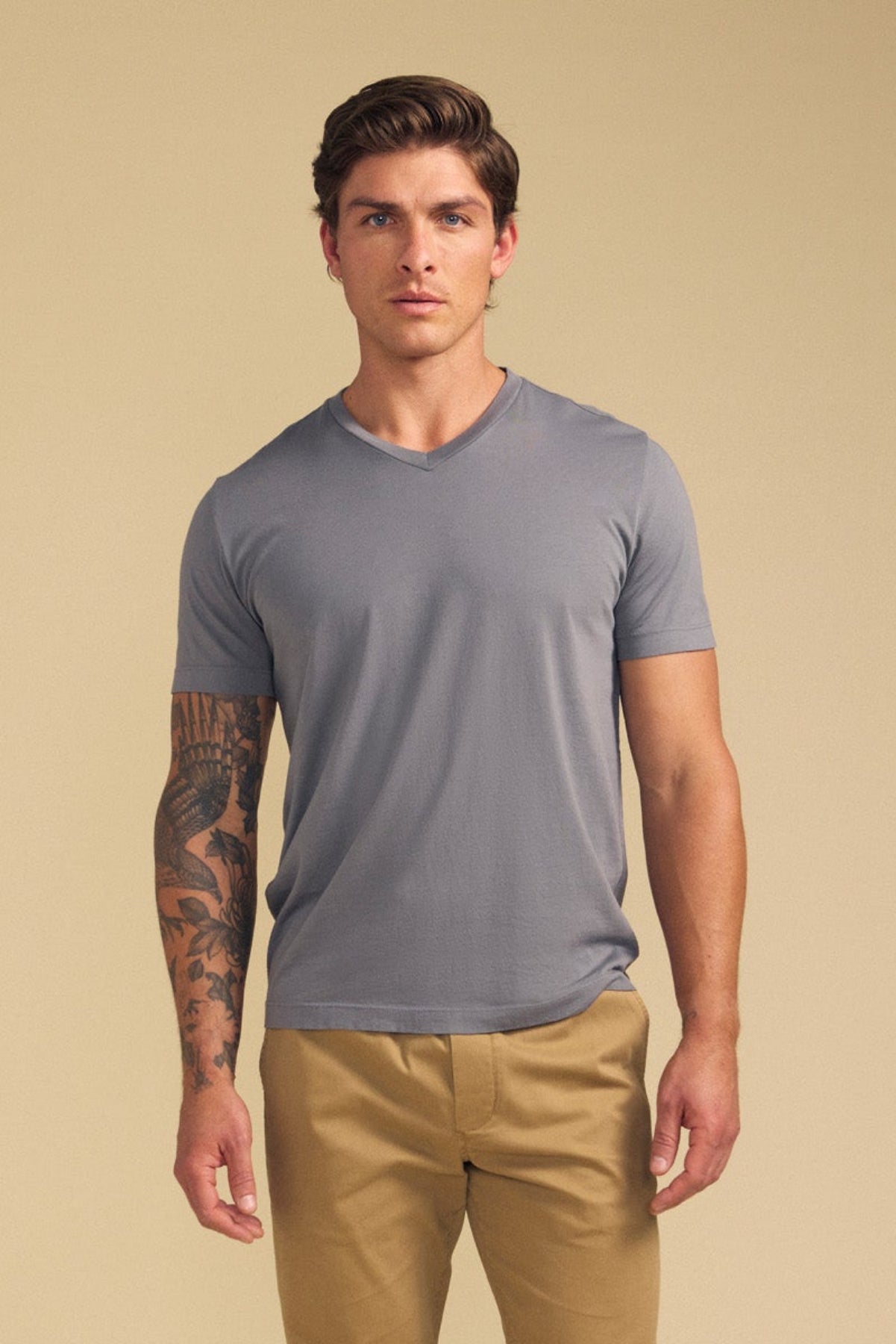 A man stands facing forward against a plain beige background, wearing the SAMSEN TEE by Velvet by Graham & Spencer in gray with a v-neckline and tan pants, showcasing timeless everyday style.