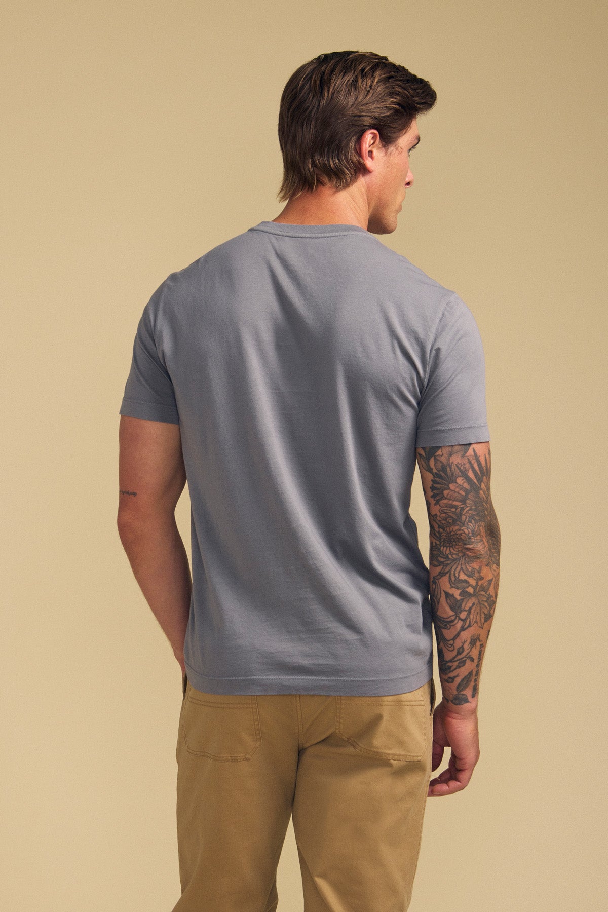 A man faces away, wearing the light gray SAMSEN TEE by Velvet by Graham & Spencer and khaki pants. An intricate black tattoo decorates his right arm. The plain beige background highlights his casual look.-41702121570497