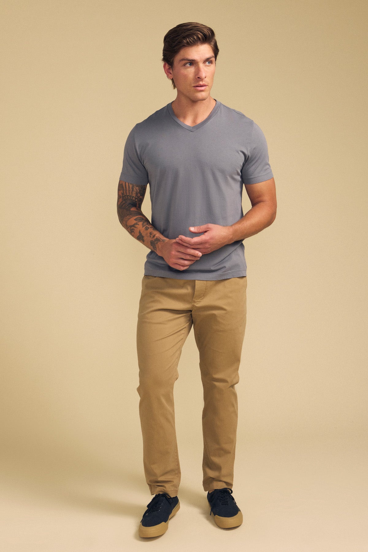 A man stands against a beige background in the SAMSEN TEE by Velvet by Graham & Spencer, a gray Pima jersey V-neck, paired with khaki pants and black sneakers with tan soles. He has a tattoo on his right forearm.