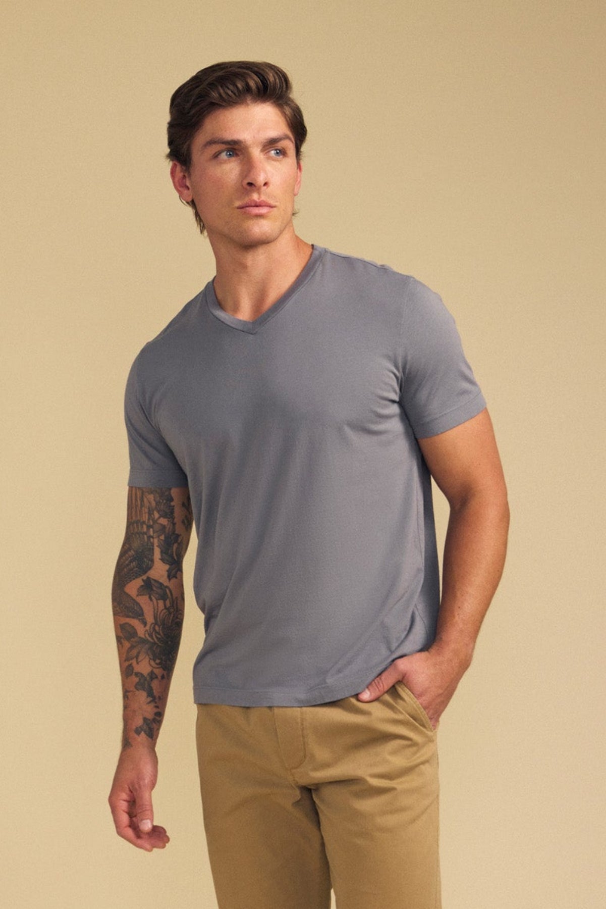 A man with short brown hair and a forearm tattoo wears the Velvet by Graham & Spencer SAMSEN TEE in gray with tan pants, standing against a beige background—a look that's perfect for effortless everyday wear.